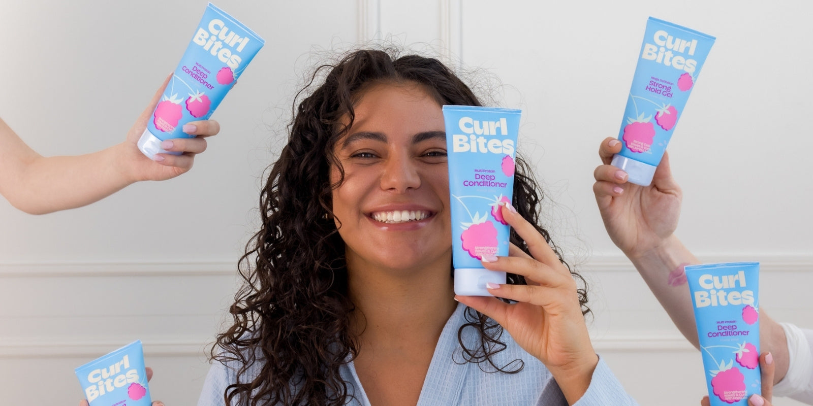Frau mit dunklen Locken lächelt und hält Curl Bites Deep Conditioner aus der Power Bites Kollektion, umgeben von weiteren Händen mit dem gleichen Produkt.