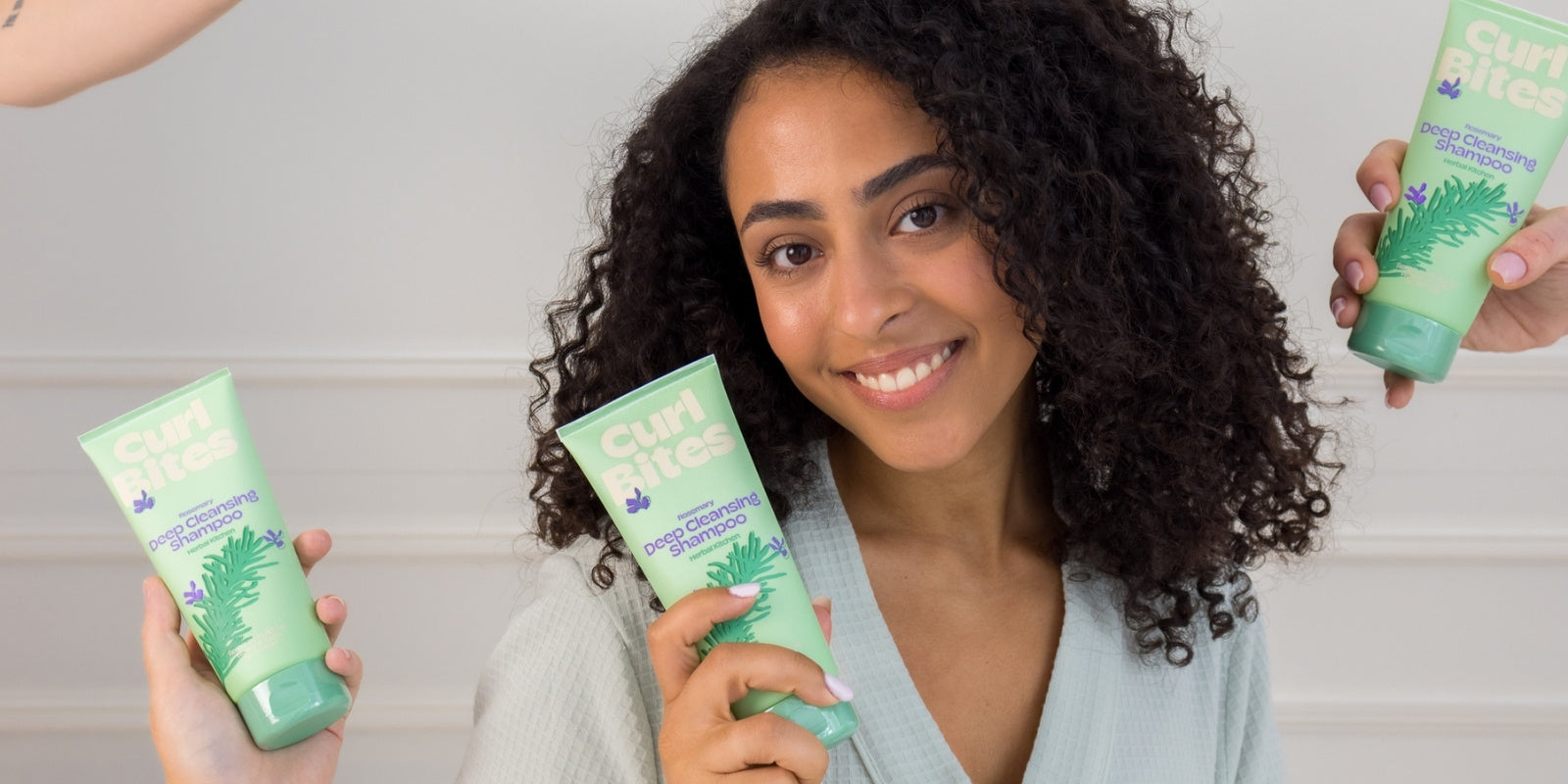 Frau mit lockigem Haar hält eine Tube Curl Bites Deep Cleansing Shampoo und lächelt, während links und rechts weitere Hände das gleiche Produkt zeigen.