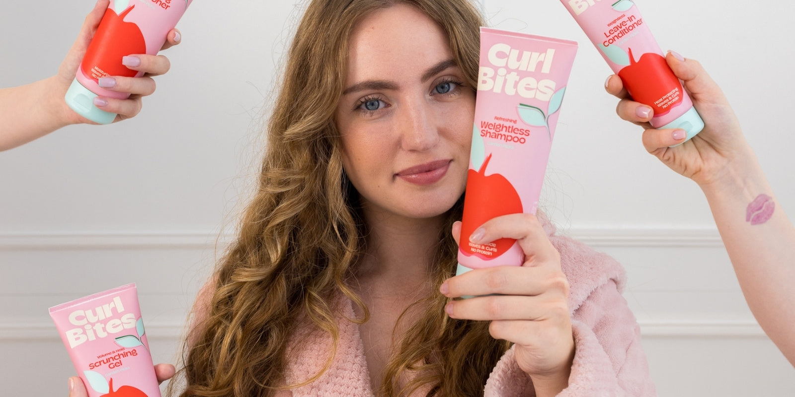 Frau mit lockigem Haar im rosa Bademantel hält ein Curl Bites Weightless Shampoo, umgeben von weiteren Händen mit Produkten der Garden Picks Kollektion