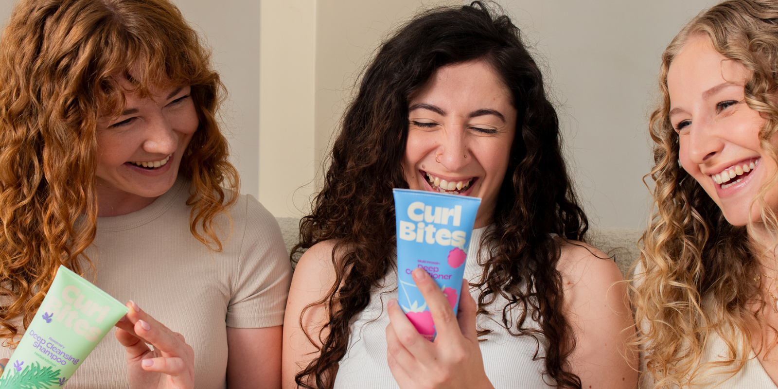 Drei lachende Frauen mit lockigem Haar sitzen nebeneinander und halten Curl Bites Haarpflegeprodukte in den Händen.