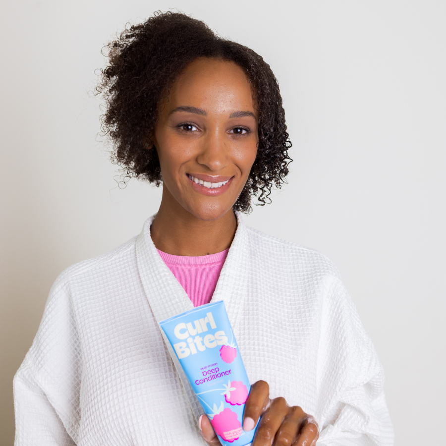 Person mit lockigem Haar und Bademantel hält eine Tube Curl Bites Deep Conditioner und lächelt in die Kamera.