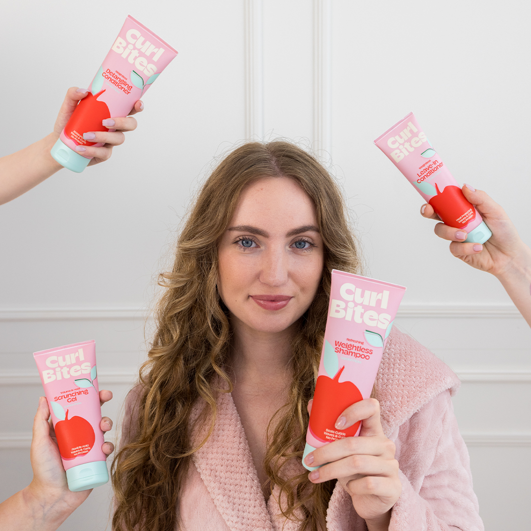 Person mit langen, lockigen Haaren hält Curl Bites Produkte – führt zur Kollektion für stärkendes Haarpflegeprogramm.