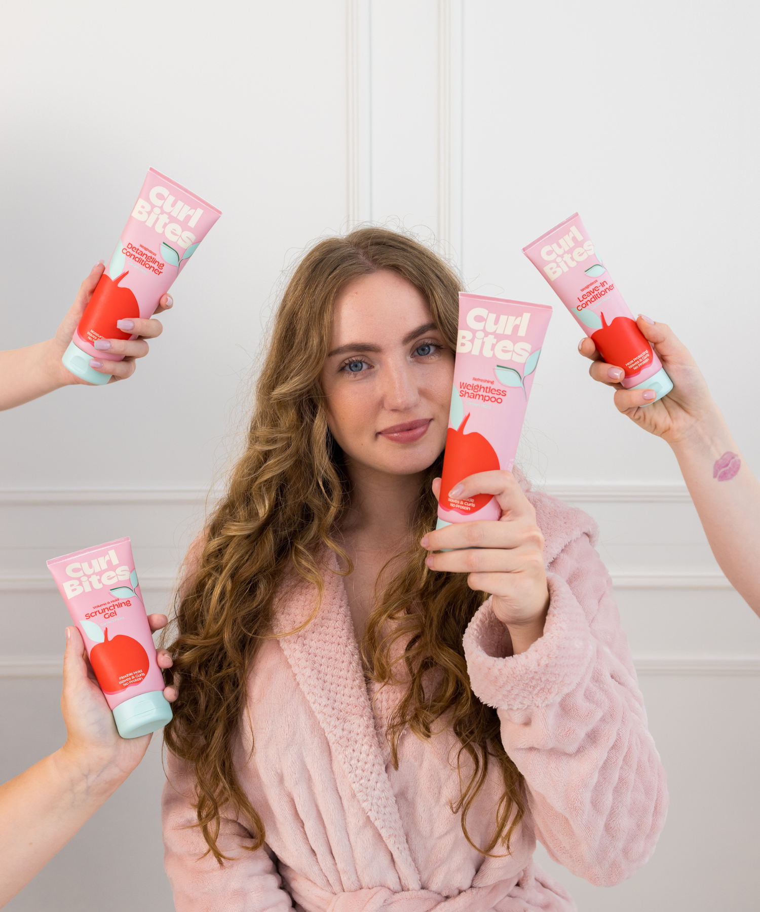 Person mit lockigem Haar in rosa Bademantel hält Curl Bites Shampoo, umgeben von weiteren Produkten – führt zur Produktreihe ‘Garden Picks’ mit schwerelosen Produkten.