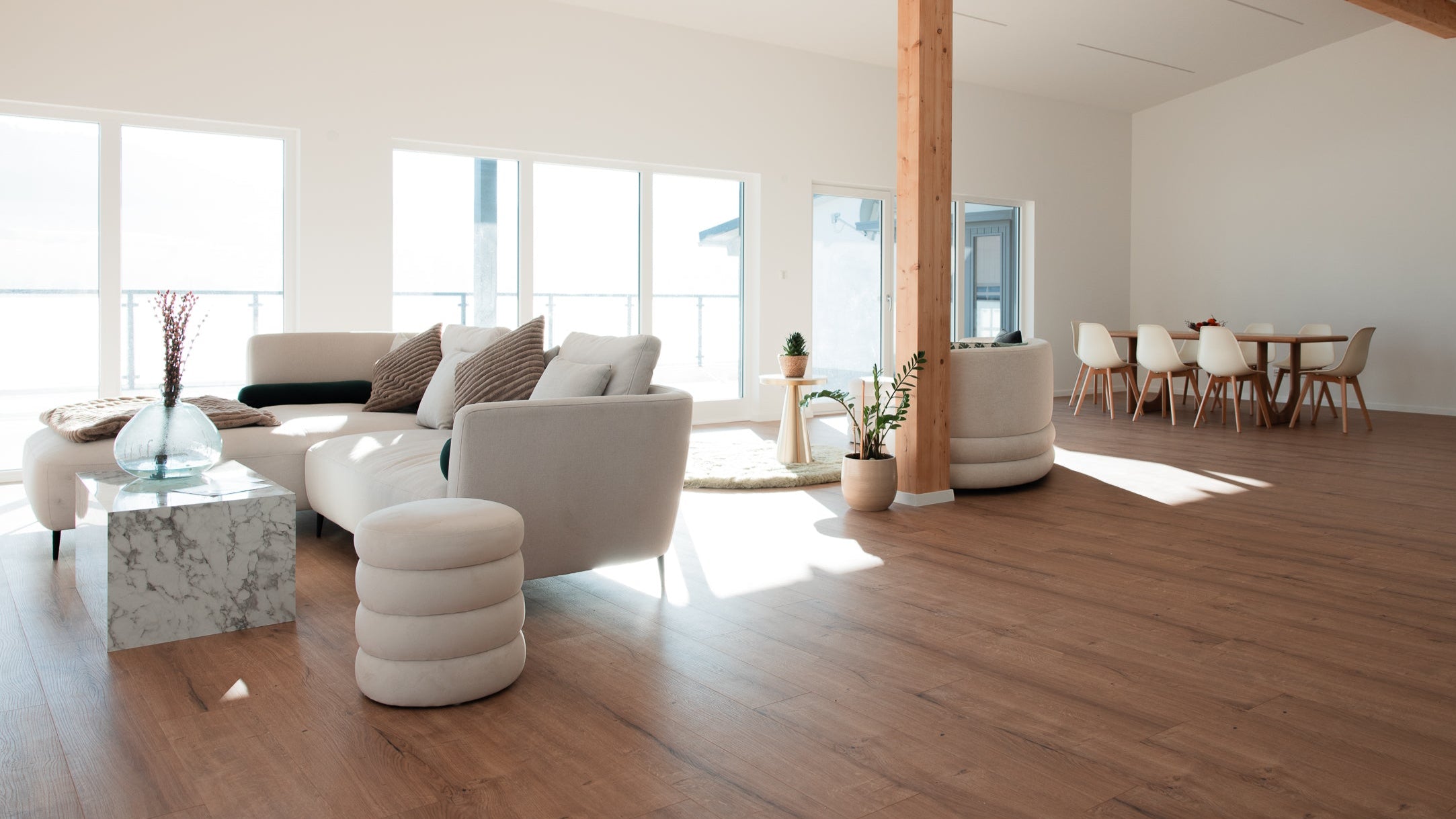 Helles, modern eingerichtetes Loft mit großen Fenstern, einem weißen Sofa mit Kissen und Decke, einem runden Hocker und einem Esstisch mit weißen Stühlen im Hintergrund. Holzboden und Holzbalken sorgen für eine warme Atmosphäre.