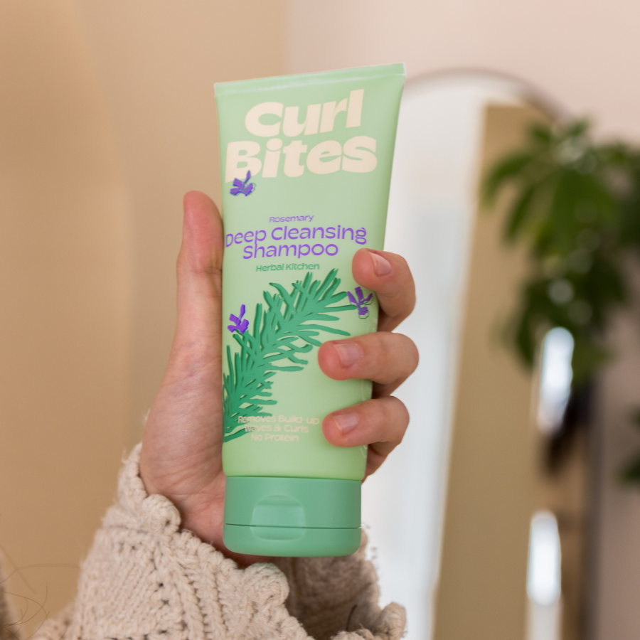 Hand hält Tube Curl Bites Deep Cleansing Shampoo mit grünem Design und Rosmarin-Illustration vor neutralem Hintergrund.