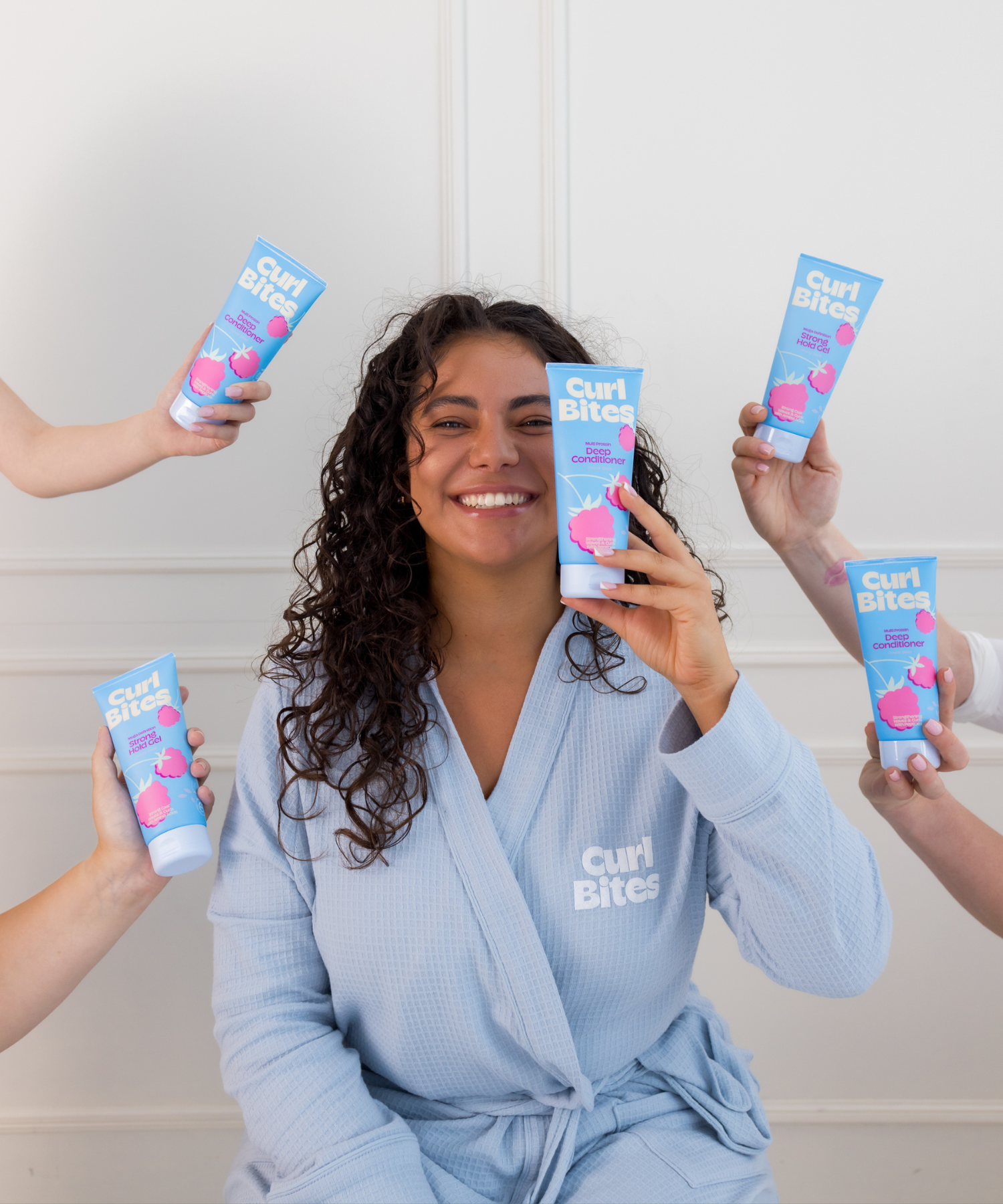Person mit lockigem Haar in blauem Bademantel hält Curl Bites Deep Conditioner – führt zur Produktreihe ‘Power Bites’ zum Haar stärken