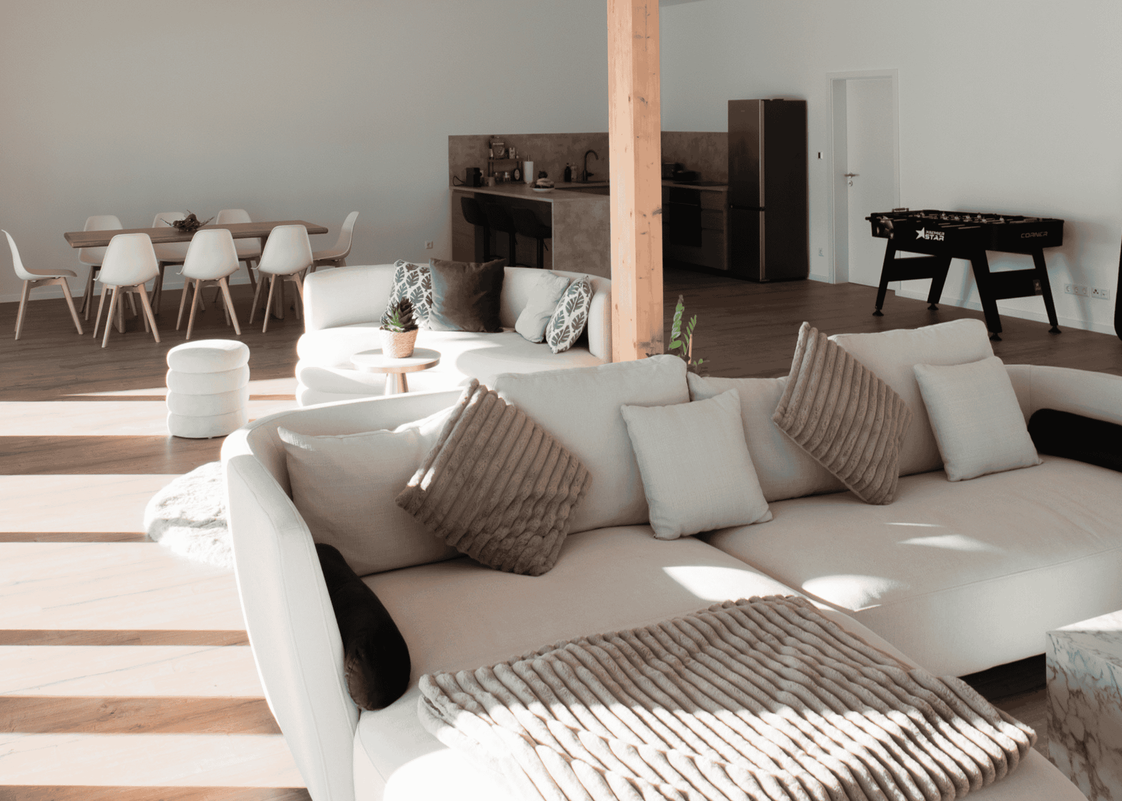 Helles Loft mit gemütlichem Wohn- und Essbereich, offener Küche, Esstisch mit weißen Stühlen, Sofas mit Kissen und Decken sowie einem Tischkicker im Hintergrund